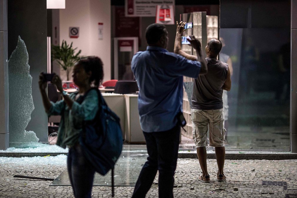 مردم در حال گرفتن عکس از در ورودی یک بانک تخریب شده توسط معترضان طی یک اعتصاب ملی در برابر لایحه اصلاح رفاه اجتماعی دولت برازیل. تصویر از AFP.